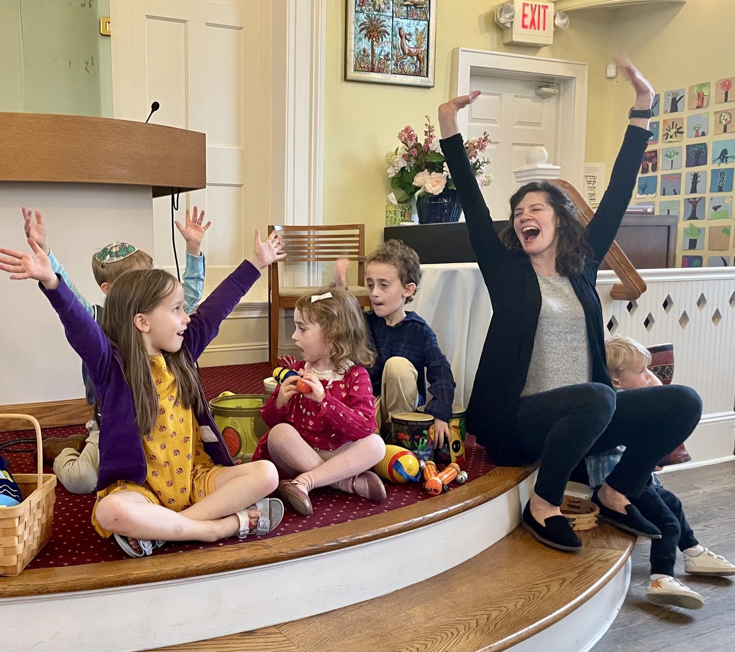 Young children sitting on the bimah steps with shakers and drums, arms in the air mid-song.