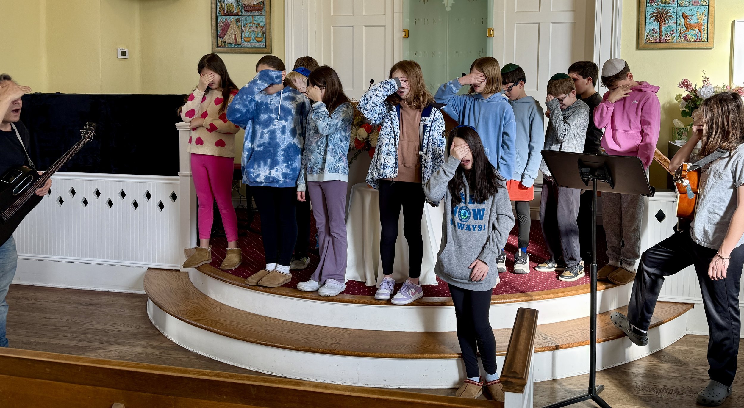 A class of students standing on the bimah with hands over their eyes for the Shema, accompanied by guitars.