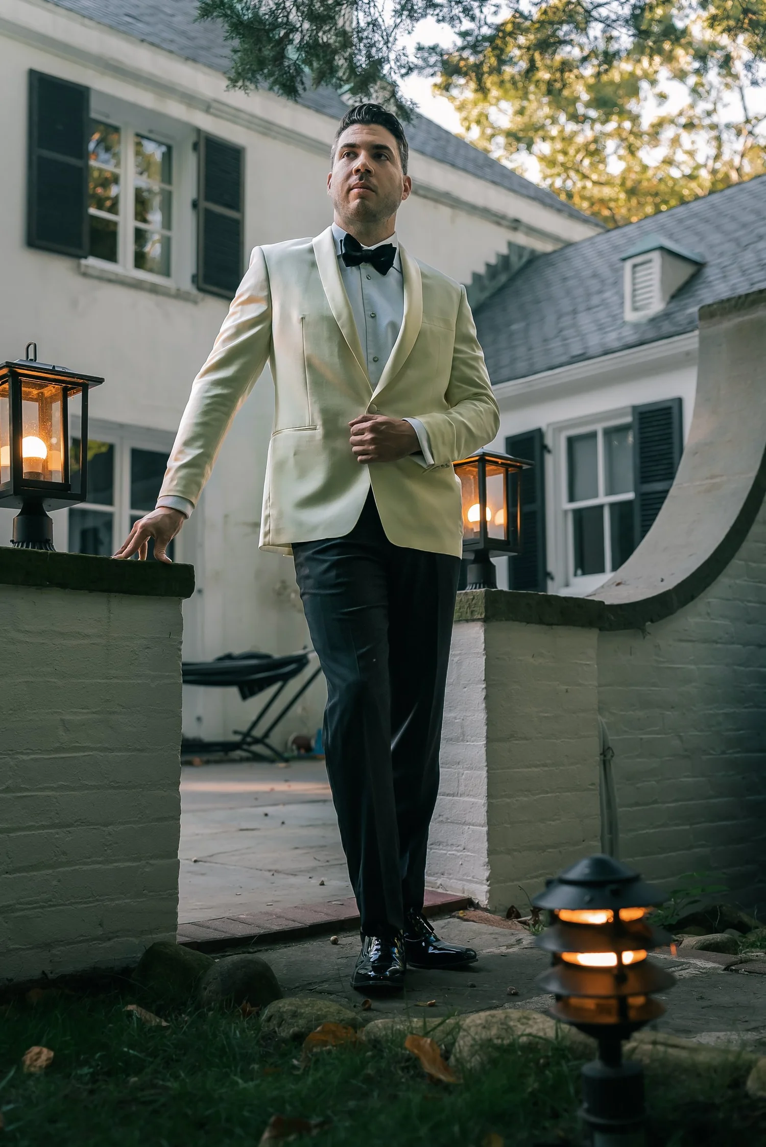 Joshua in an ivory dinner jacket and black bow tie walks through a garden gate between two lit lanterns.