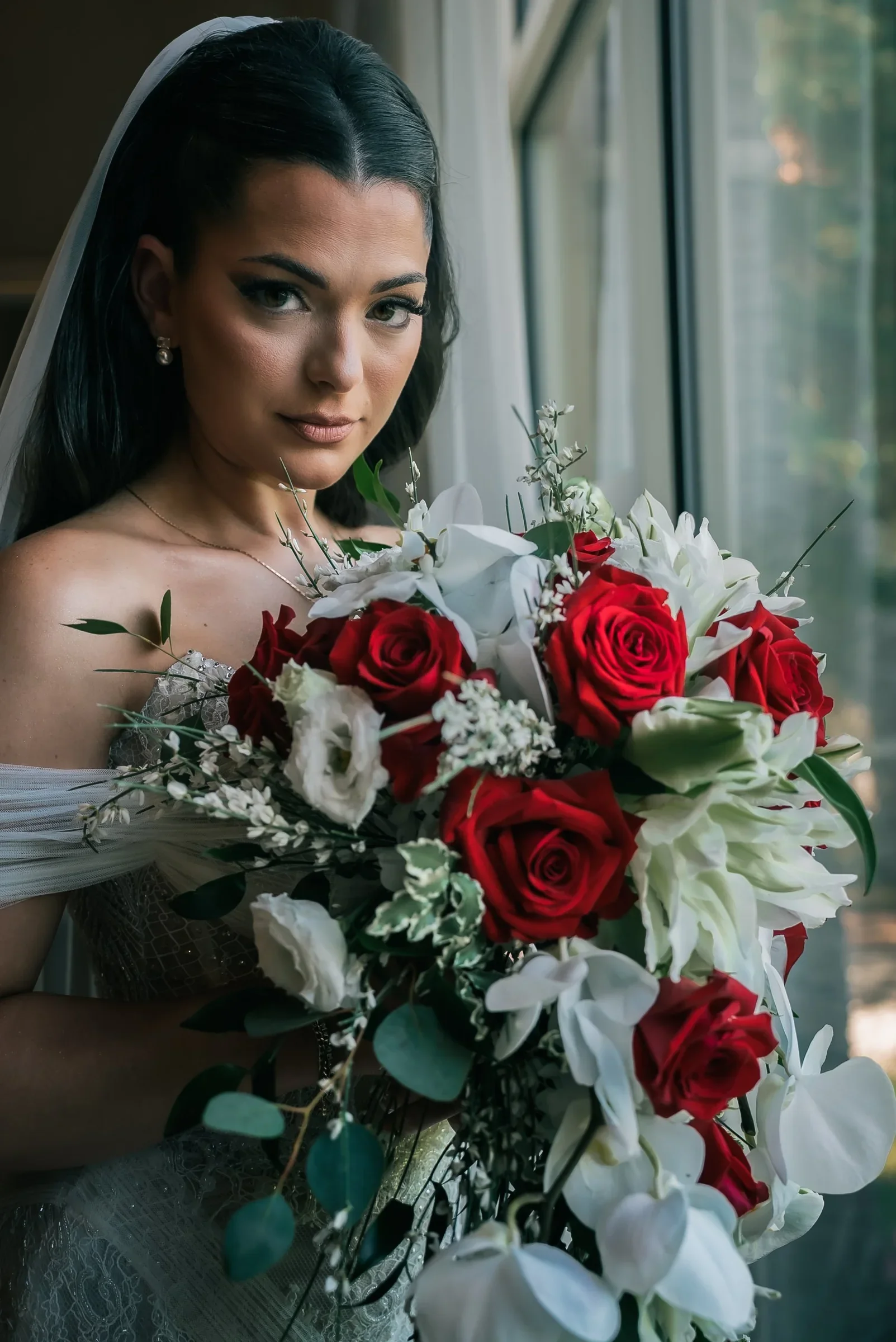 Vanessa holding a cascading bouquet of red roses and white orchids beside a tall window
