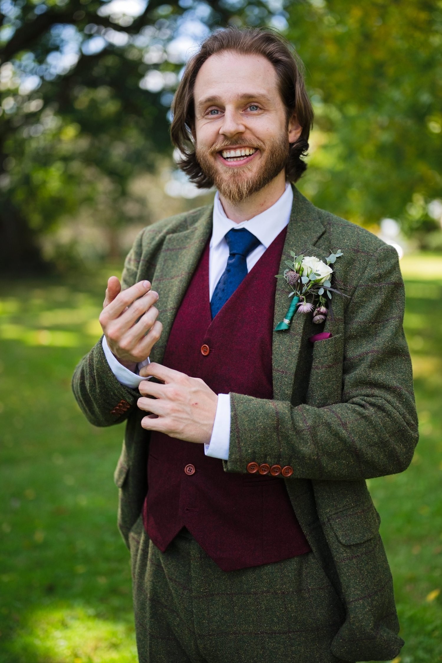 Jason grinning on the lawn in a green tweed suit, burgundy waistcoat and blue tie, with a white rose on his lapel