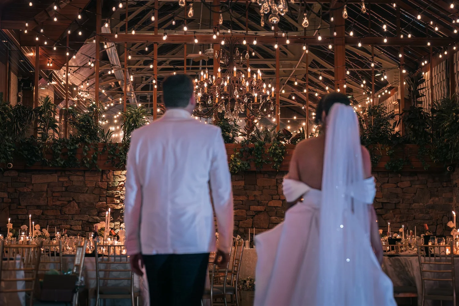 Patricia and Joshua look across The Somerley reception room beneath chandeliers and string lights.