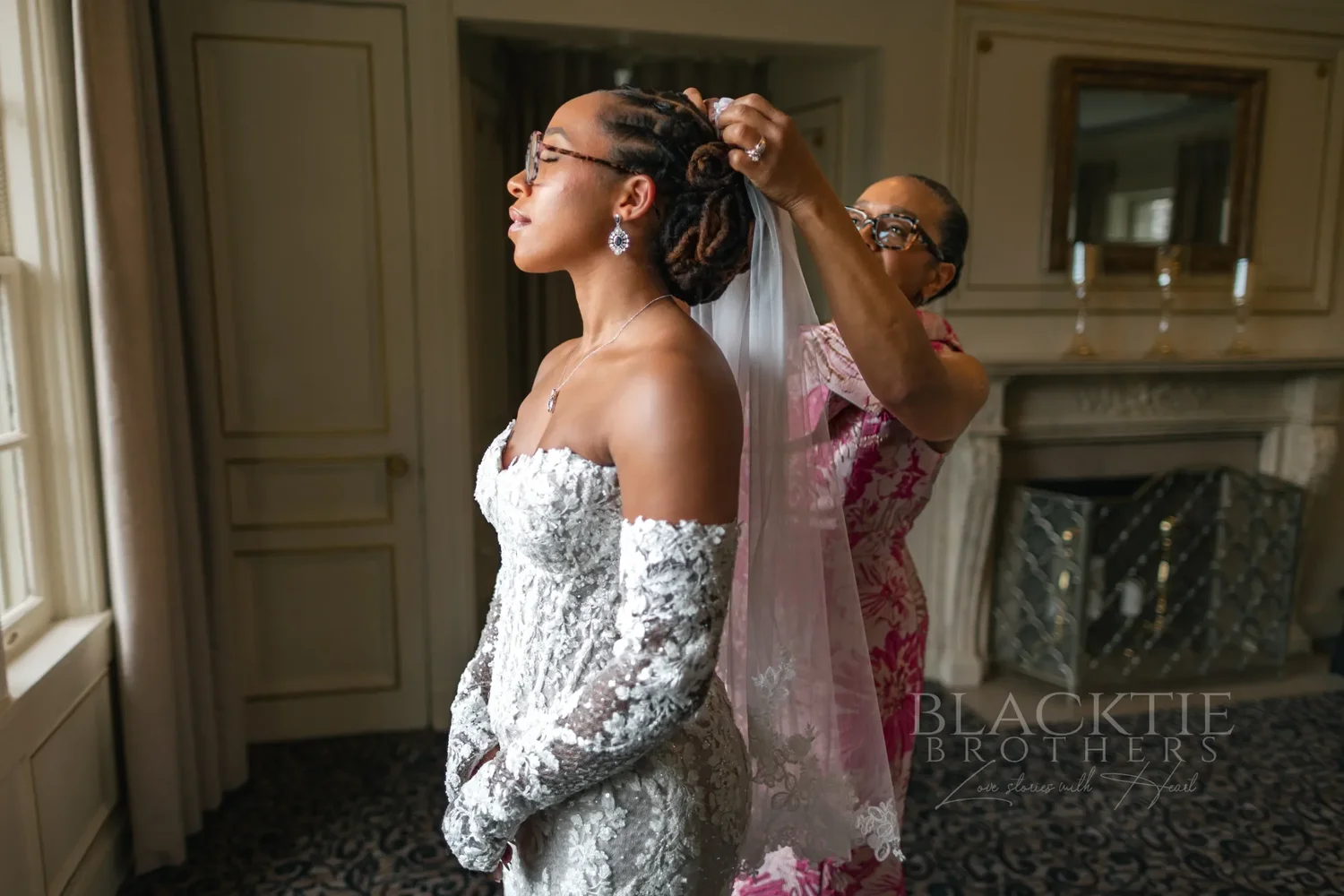 A woman in a pink floral dress settling the bride’s veil in a paneled room with a fireplace