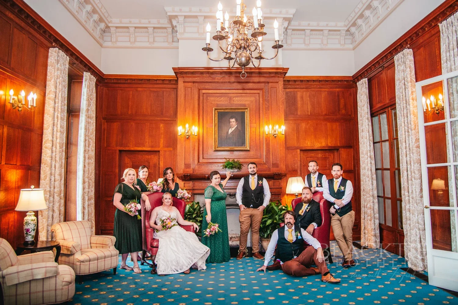 Amanda and Chris with their wedding party in a wood-paneled room with a chandelier at The Otesaga