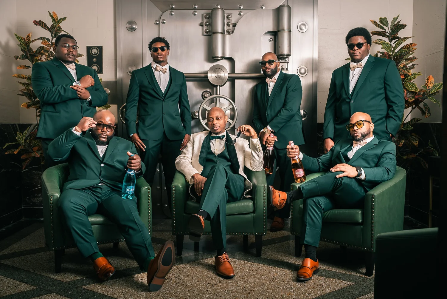 David in an ivory jacket sits with his groomsmen in green suits and sunglasses in front of a large steel vault door.