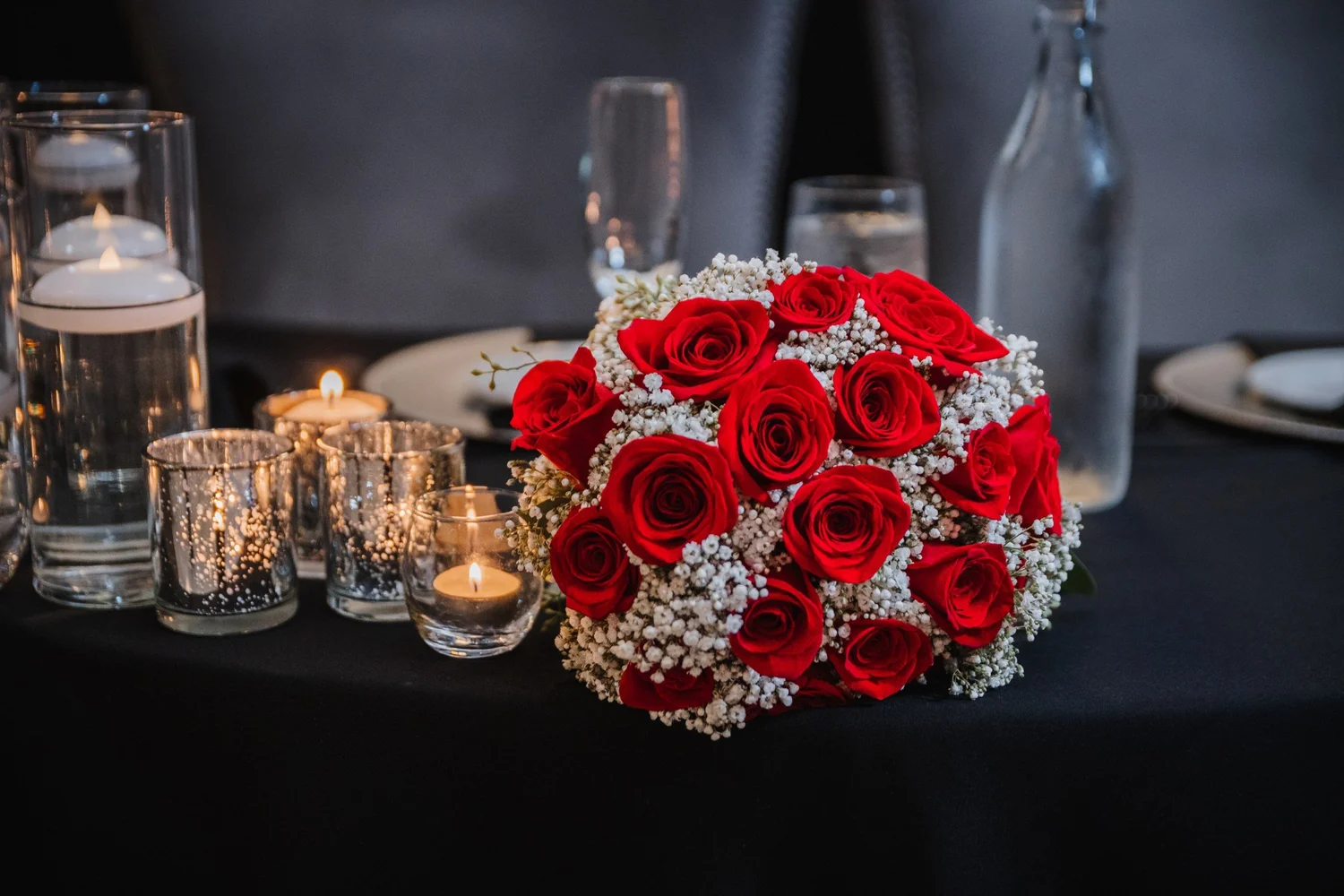 A bouquet of red roses and baby’s breath on a black tablecloth beside floating candles
