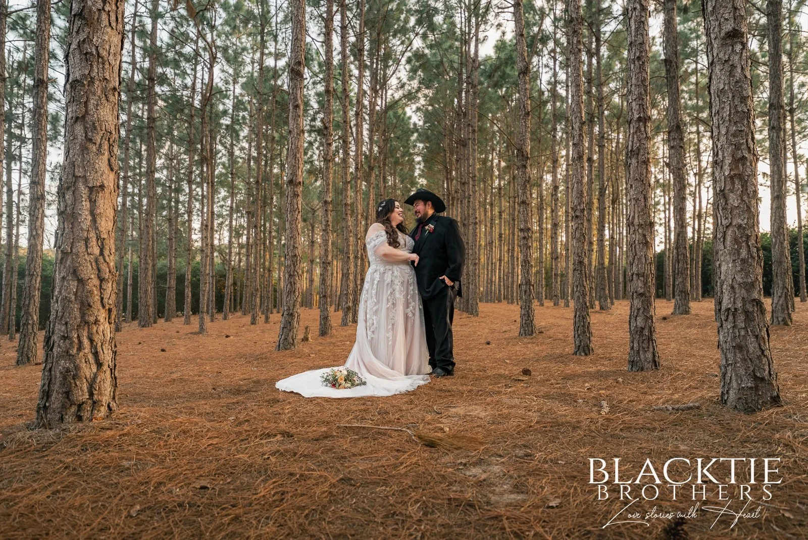 Alyssa and Alberto smile at each other among tall pine trees, with her wedding gown train spread on the pine-covered ground.
