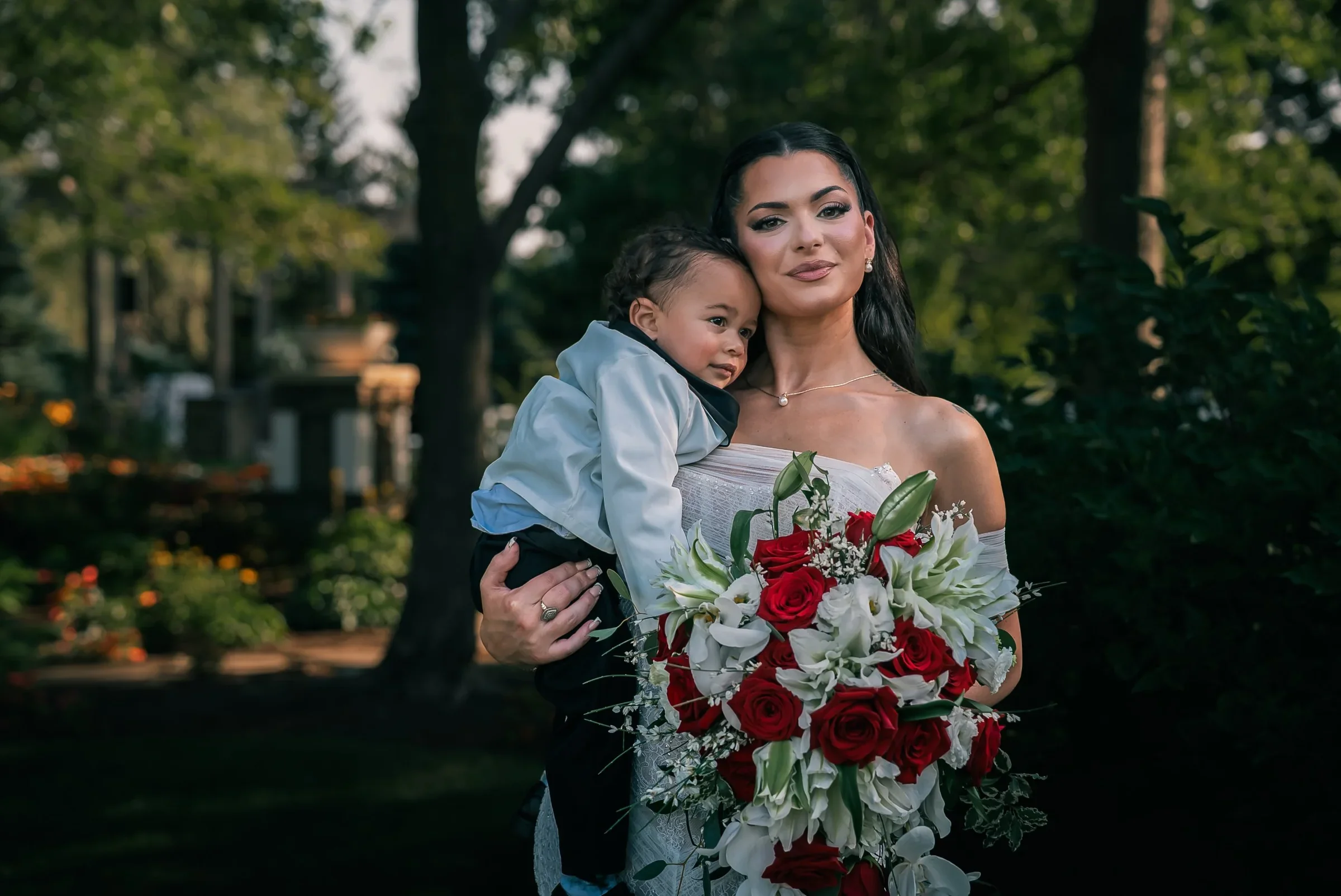 Vanessa holding a sleepy toddler in a white tuxedo jacket against her cheek, her bouquet in the other arm