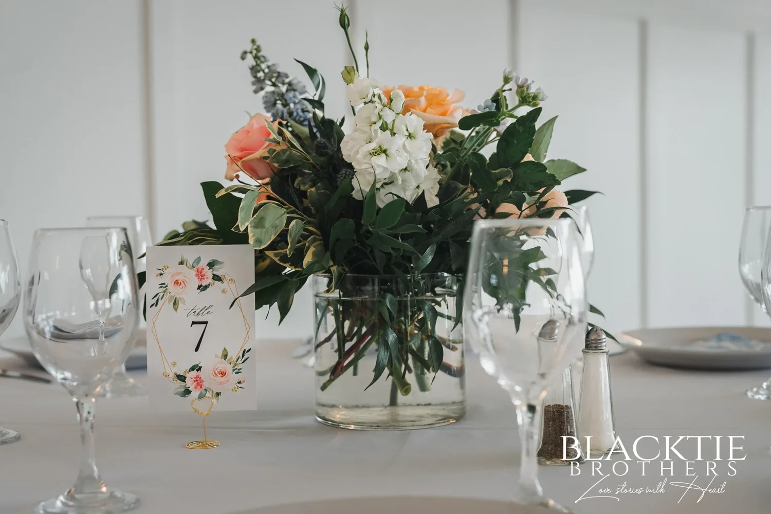 A reception table set with wine glasses, a floral table number card and a centerpiece of peach roses, white stock and greenery in a glass vase