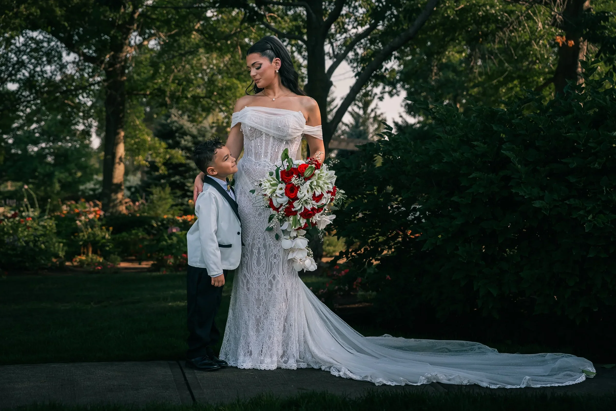 Vanessa with her long lace train spread along the garden path while a young boy in a white tuxedo jacket smiles up at her