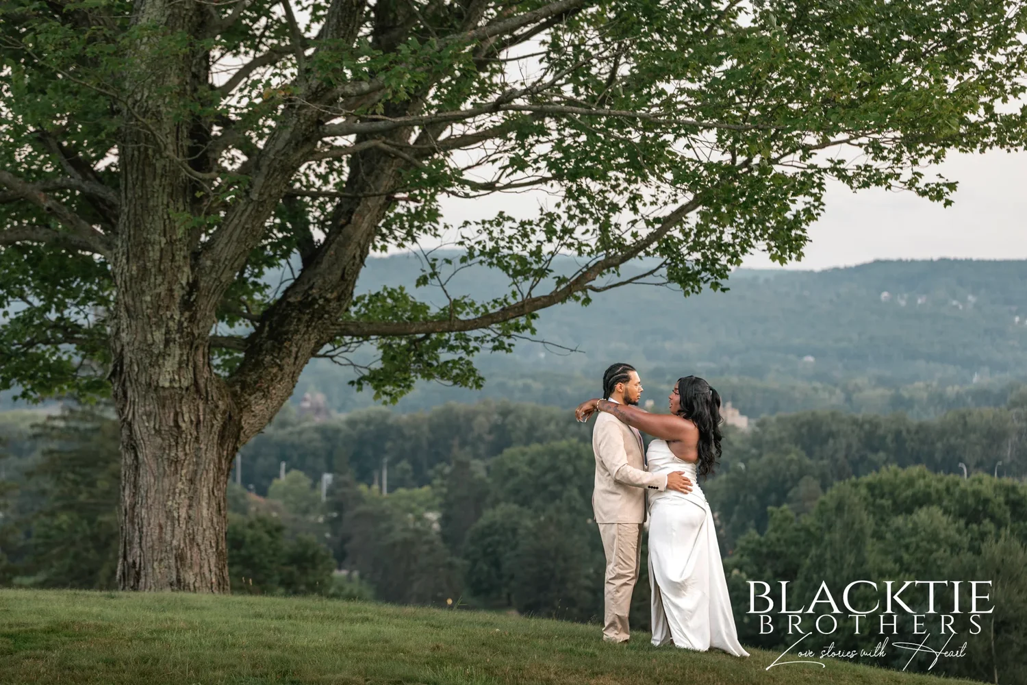A bride and groom embracing on the hill at Traditions at the Glen under the branches of a large tree, with the valley and ridge stretching out behind them