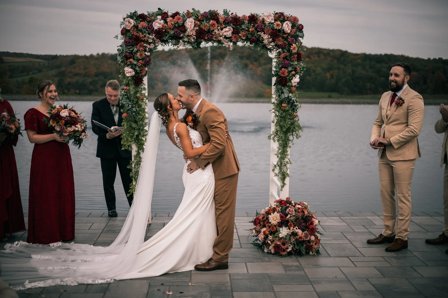 Heather and Vincent’s first kiss under the floral arch, with the pond, the fountain and the wedding party around them