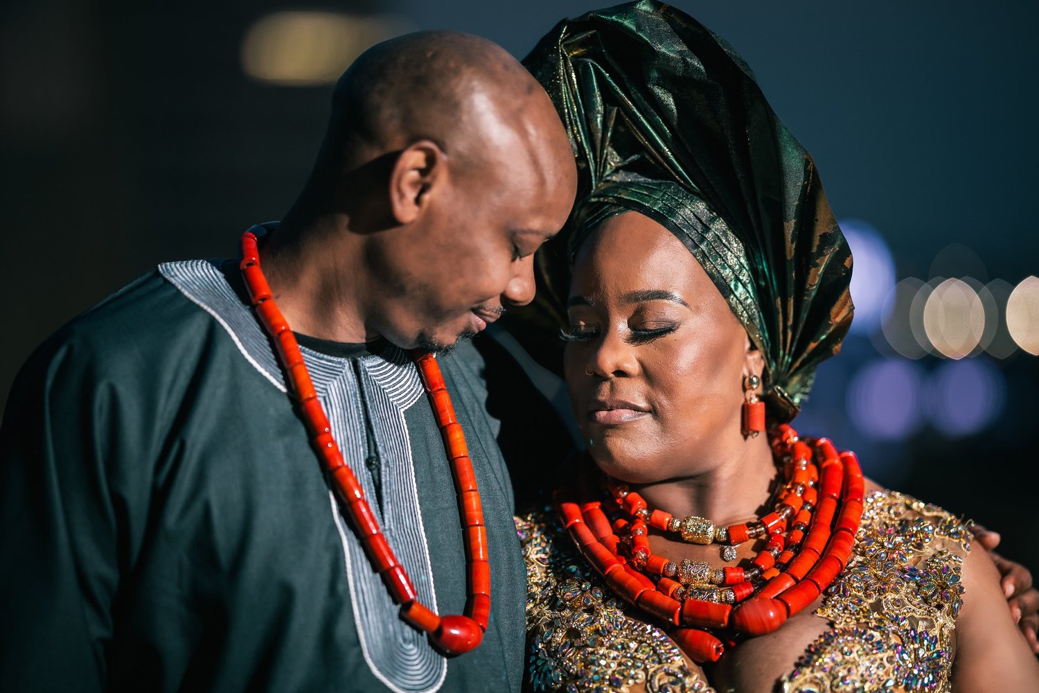 Tiffany and David rest their heads together in traditional attire and coral bead necklaces, Tiffany in a green and gold head wrap.