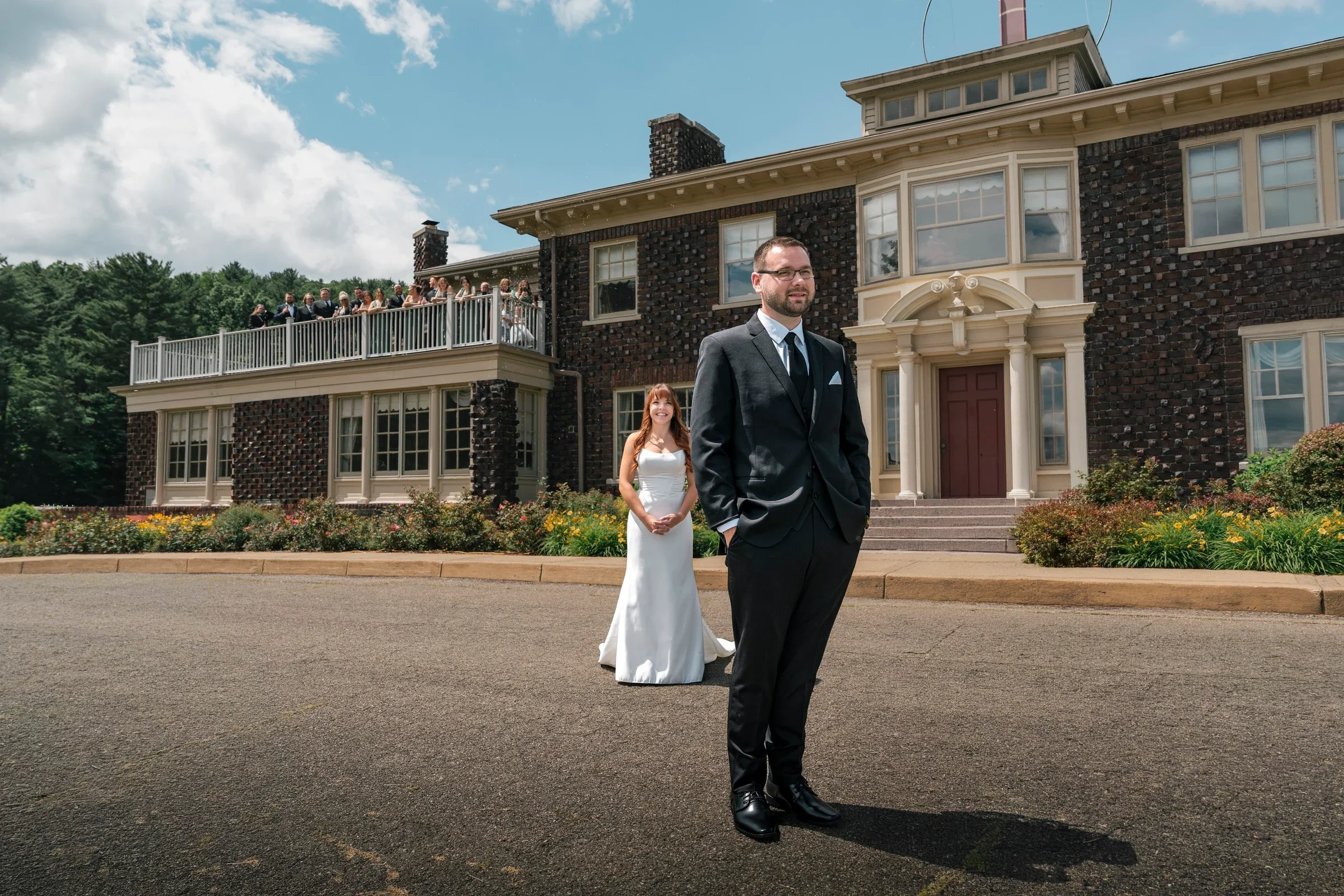 Zach waiting in the front drive of the mansion at Traditions at the Glen as Danielle walks up behind him for their first look, bridesmaids watching from the balcony