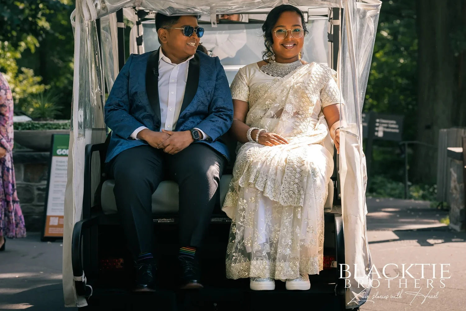Hima and Alina sitting side by side in the back of a white golf cart on a garden path, one in a blue tuxedo jacket and sunglasses, one in an embroidered ivory outfit and silver jewelry