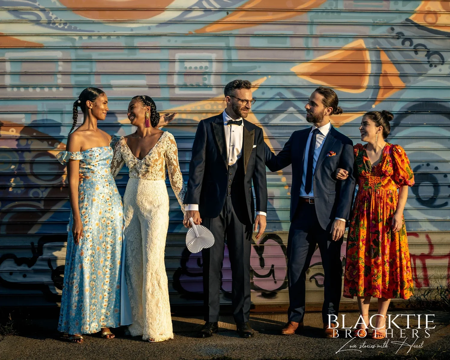 Ciara and Drew with three members of their wedding party in front of a painted roll-down gate, one laughing and one holding a white fan