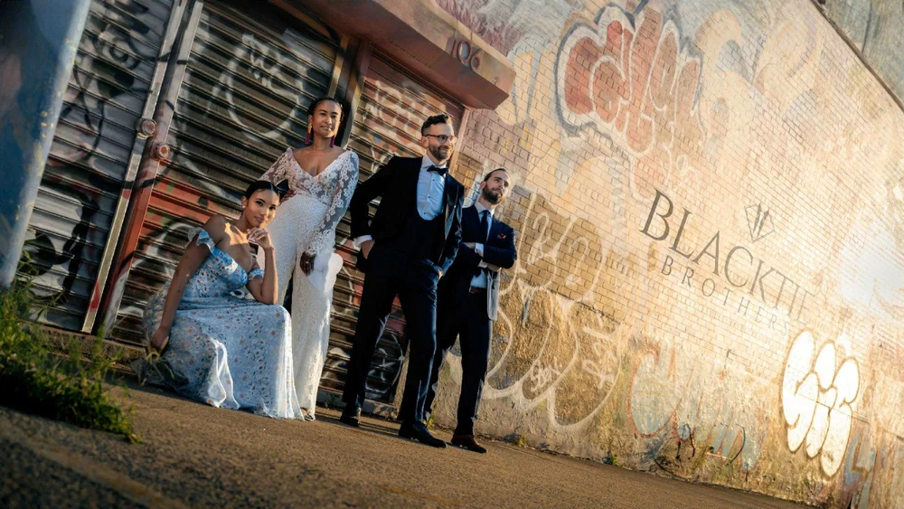 Four people in wedding attire posing beside a graffiti-covered brick wall and metal shutter.