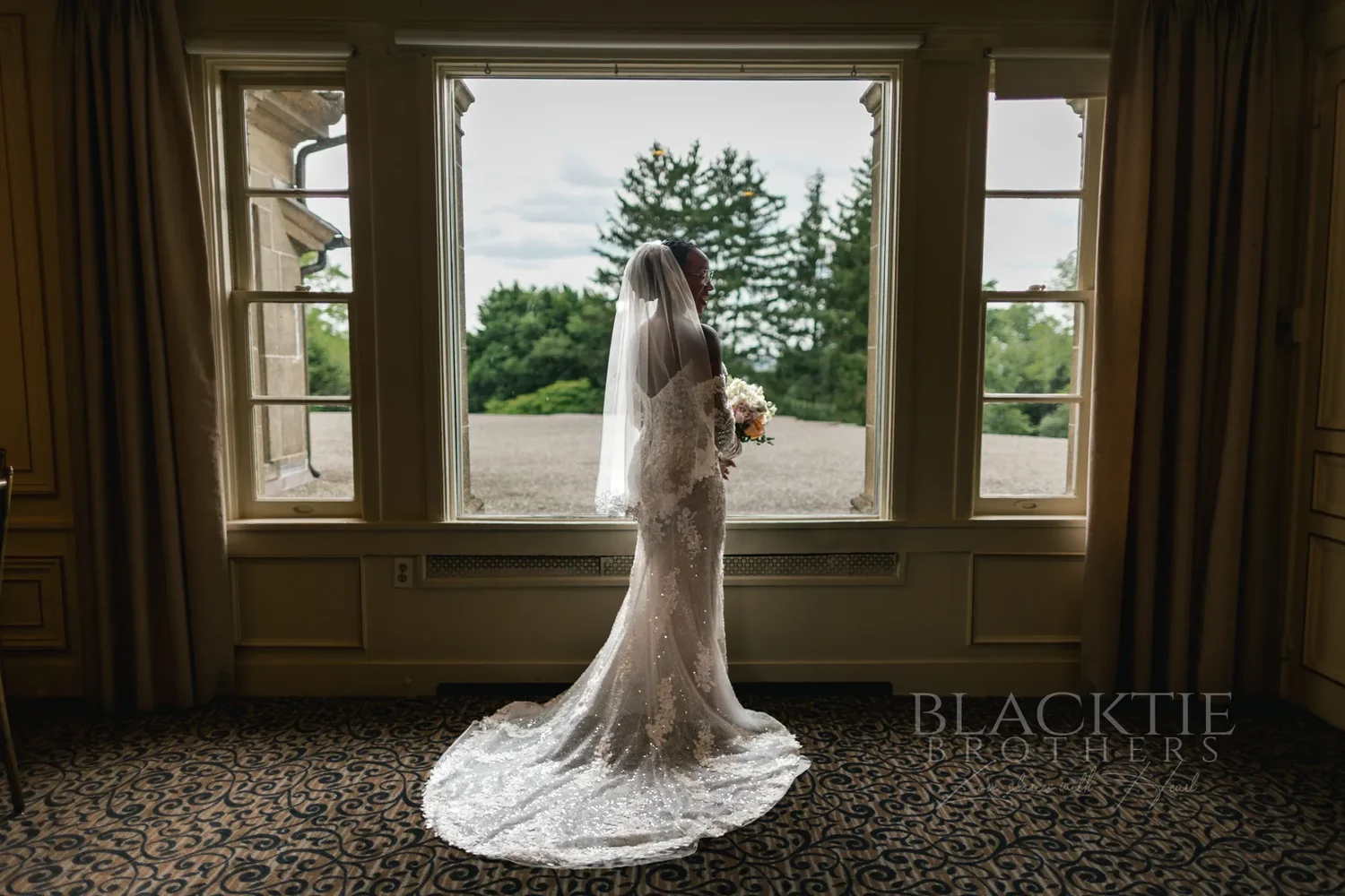 The bride in her lace gown and veil standing at a tall window in a paneled room, looking out at the trees