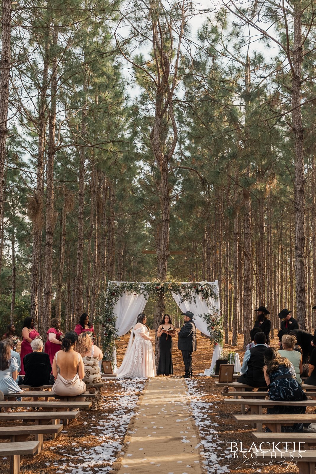 Alyssa and Alberto stand beneath a white-draped floral arch as guests watch from wooden benches under tall pine trees.