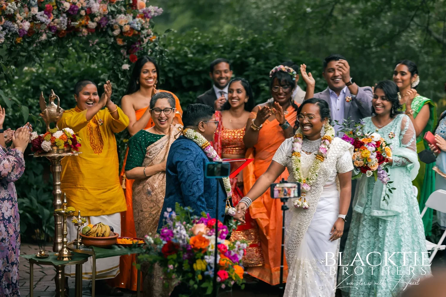 Hima and Alina in their flower garlands surrounded by cheering, clapping guests in bright saris and suits under the floral arch after the ceremony