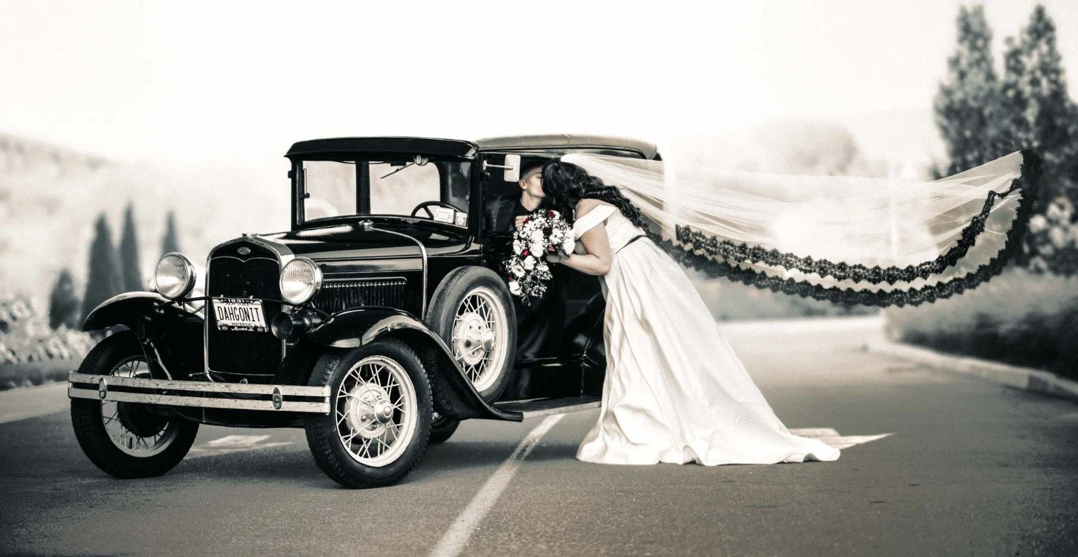 A wedding couple kisses beside a vintage car while a long, dark-trimmed veil stretches behind them.