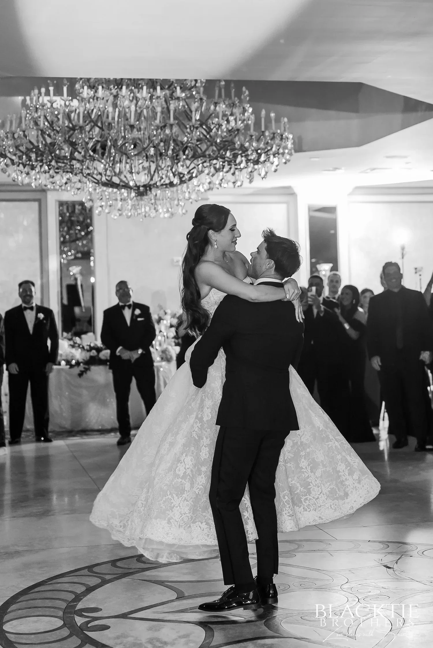 Black and white photograph of Nicholas lifting Leah during their first dance on an inlaid marble dance floor under a large crystal chandelier, guests watching around the room