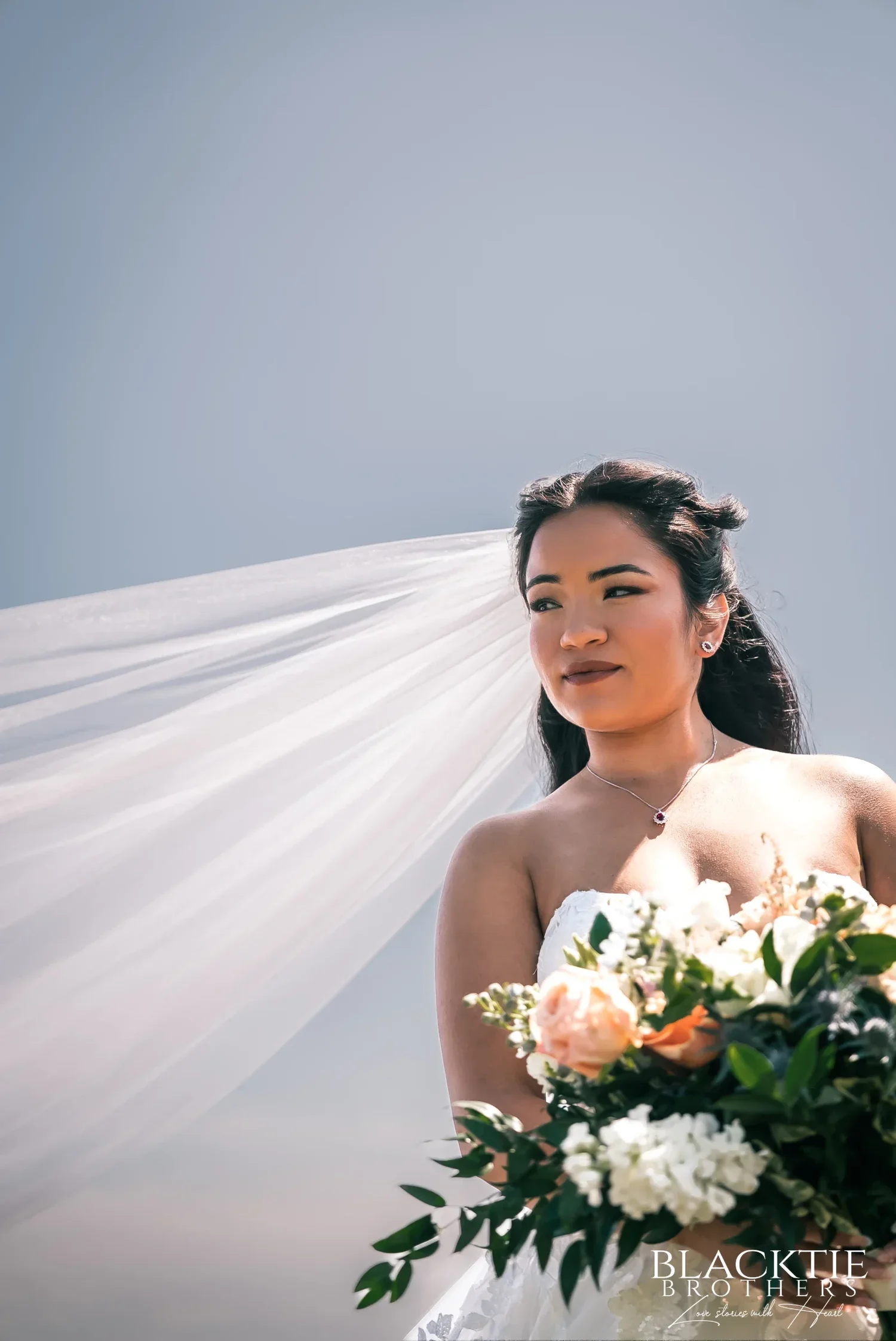 Carolina holding her bouquet of peach and white flowers by the water while the wind lifts her long veil out to the side