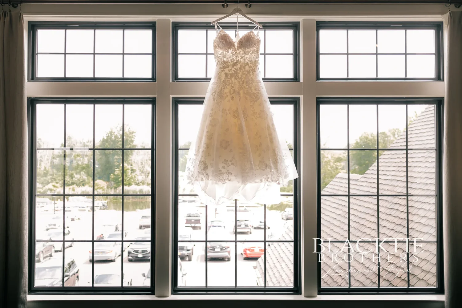 Taylor's lace wedding dress hanging in the tall window of the upstairs suite at Timber Banks, with the parking lot and pond beyond the glass