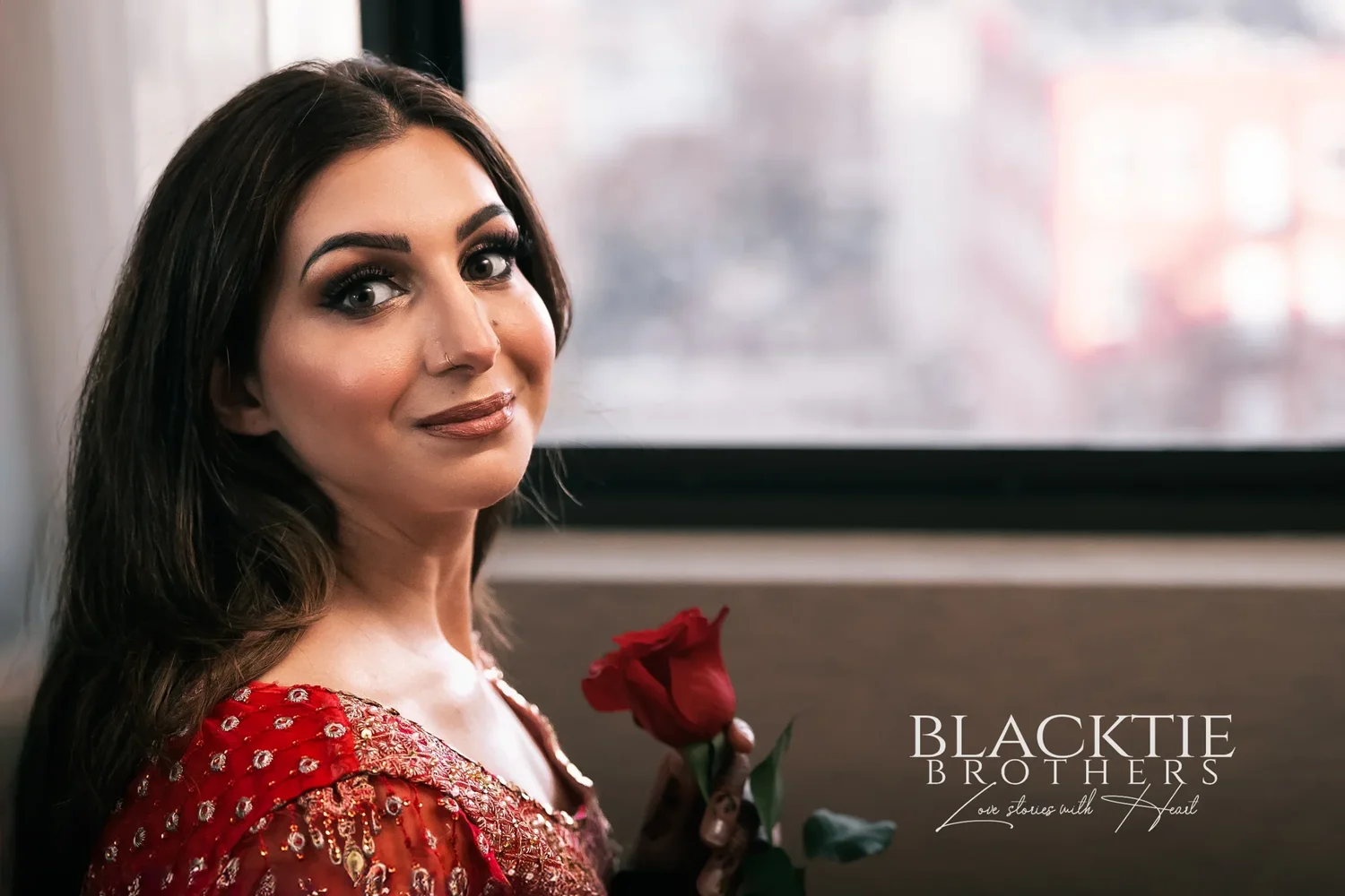 Jia in a red embroidered outfit smiling over her shoulder beside a hotel room window, holding a red rose, with downtown buildings blurred behind her