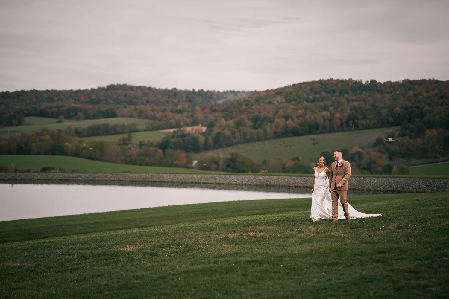 Heather and Vincent standing small on the wide lawn, the pond and the hills of the Endless Mountains stretching out behind them