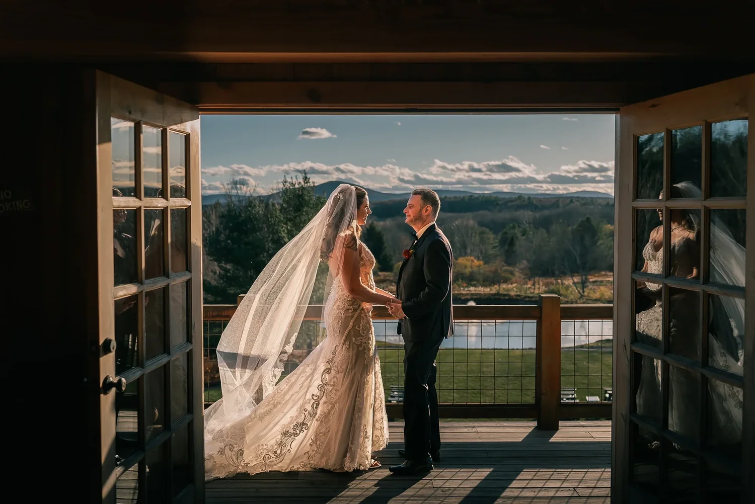 Charissa and Andrew holding hands on the Barn balcony, framed by its open doors, her long veil trailing behind her, with the lawn, the pond and the Catskills beyond