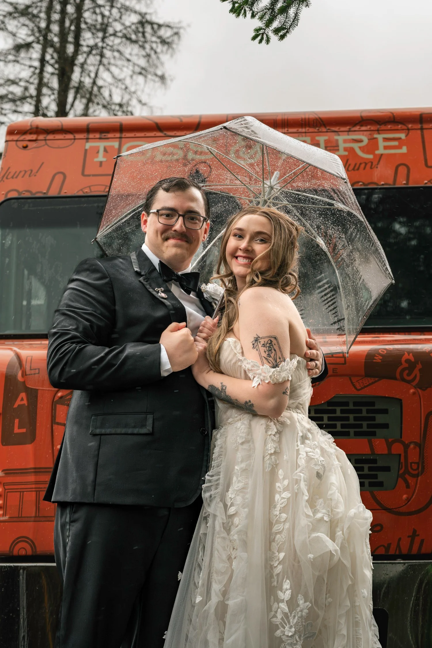 Lindsey and Noah sharing a clear umbrella in front of a vintage red fire truck