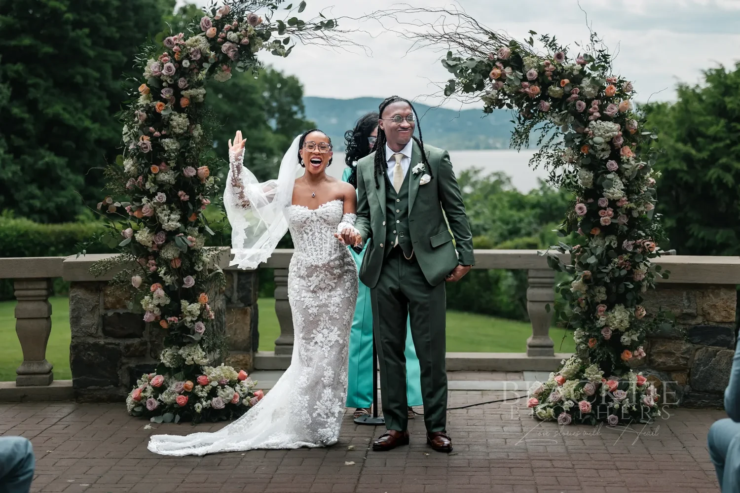 The bride raising her arm in celebration as she and the groom walk away from the floral arches, the river in the distance
