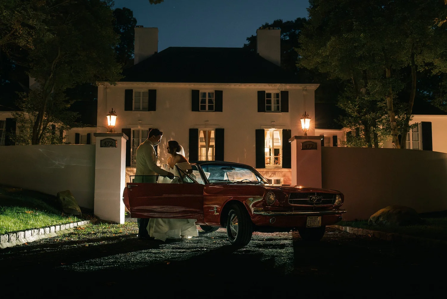 At night, Joshua holds open the door of a red vintage Mustang for Patricia in front of a white house with glowing lanterns.