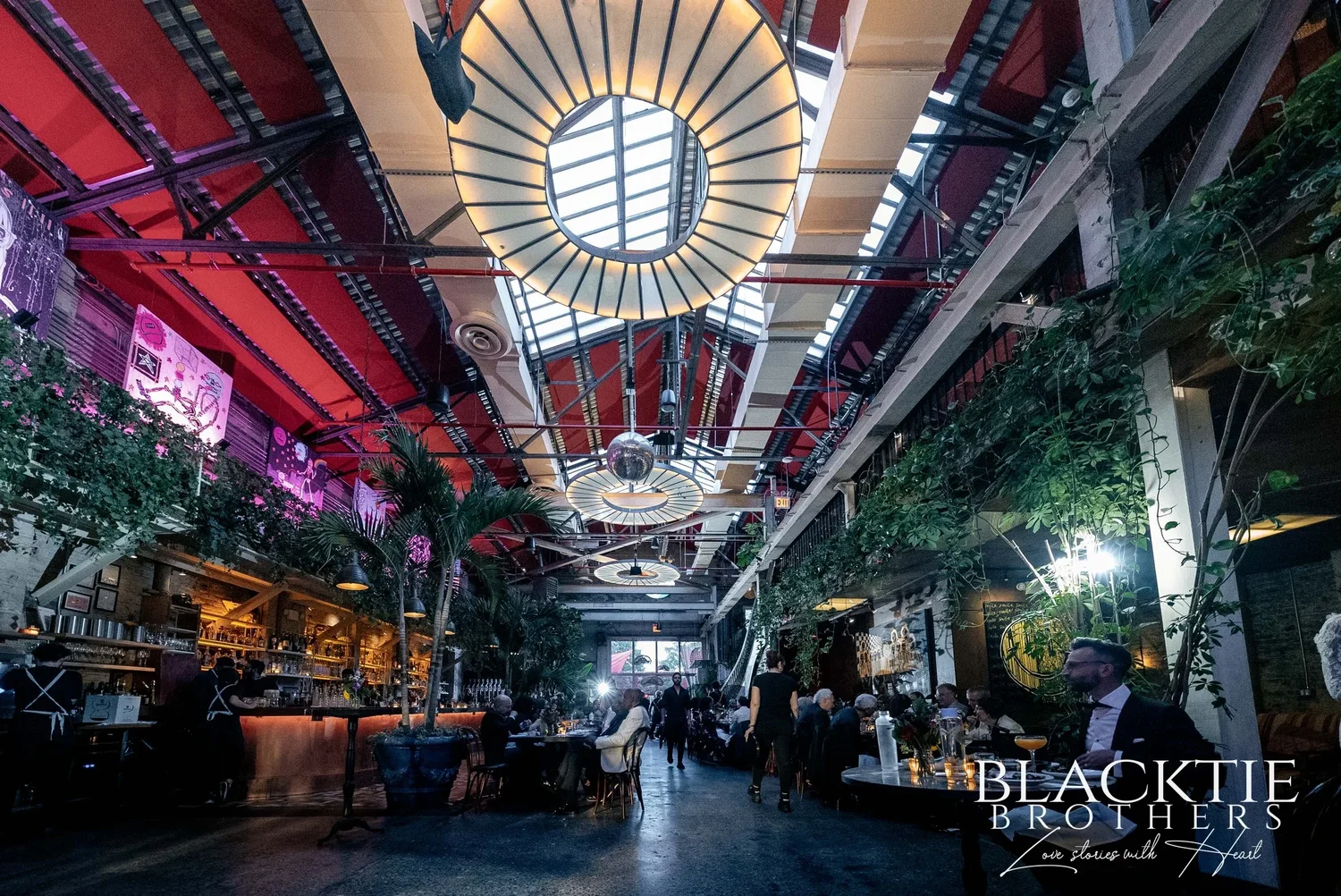 The main dining room at Fandi Mata: a long hall under a round glass skylight ringed with lights, red-painted steel trusses, round hanging lamps, plants trailing from the mezzanine and guests at candlelit tables