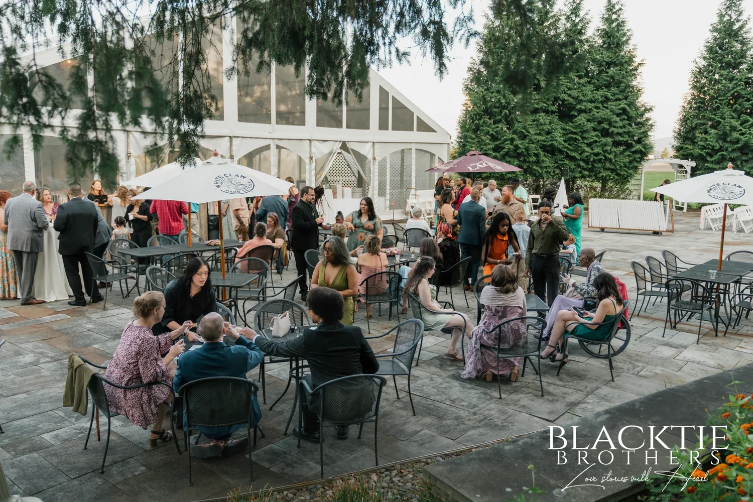 Guests at cocktail hour on the stone patio beside the white reception tent at Traditions at the Glen, with patio umbrellas and tall evergreens