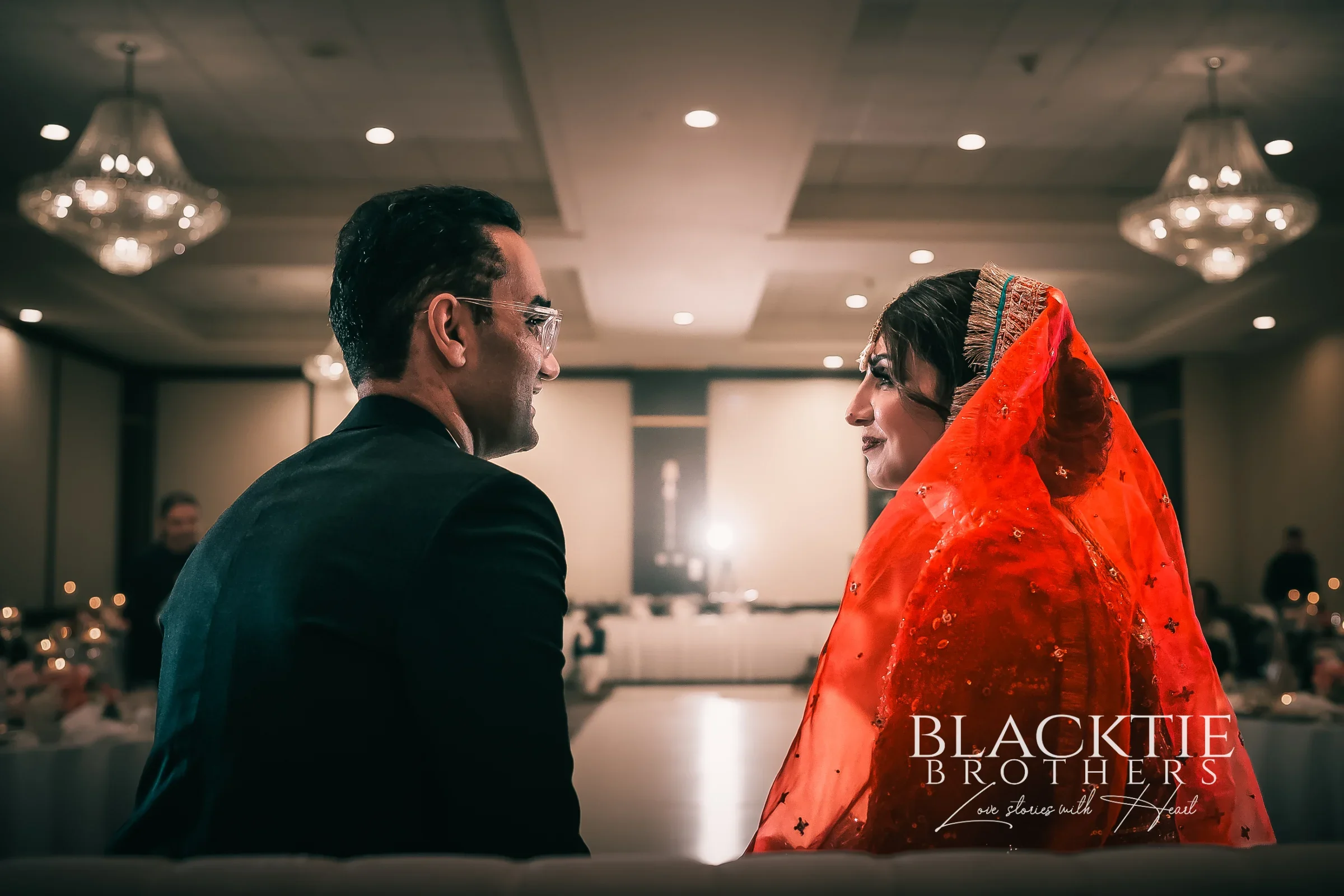 Jia and Rizwan seated on the stage at their Nikkah, turned toward each other, with the crystal chandeliers and coffered ceiling of the ballroom at the DoubleTree by Hilton Binghamton behind them