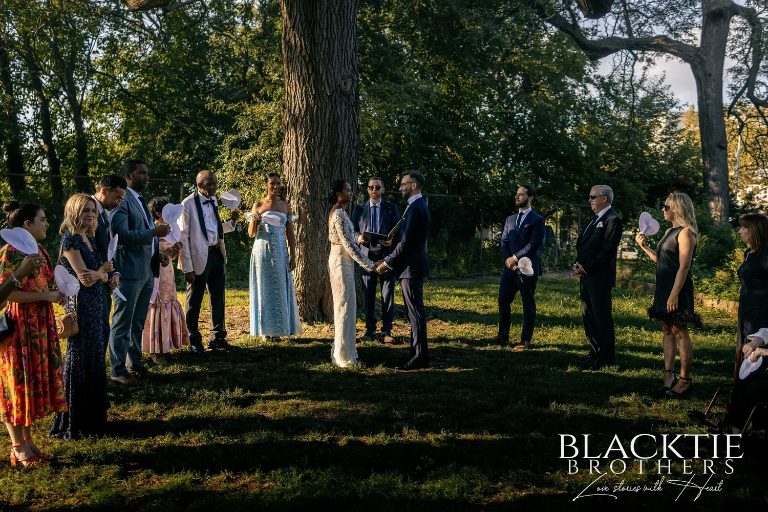 The ceremony in McCarren Park seen from across the lawn: guests standing in a wide circle on the grass around Ciara, Drew and the officiant under a large tree