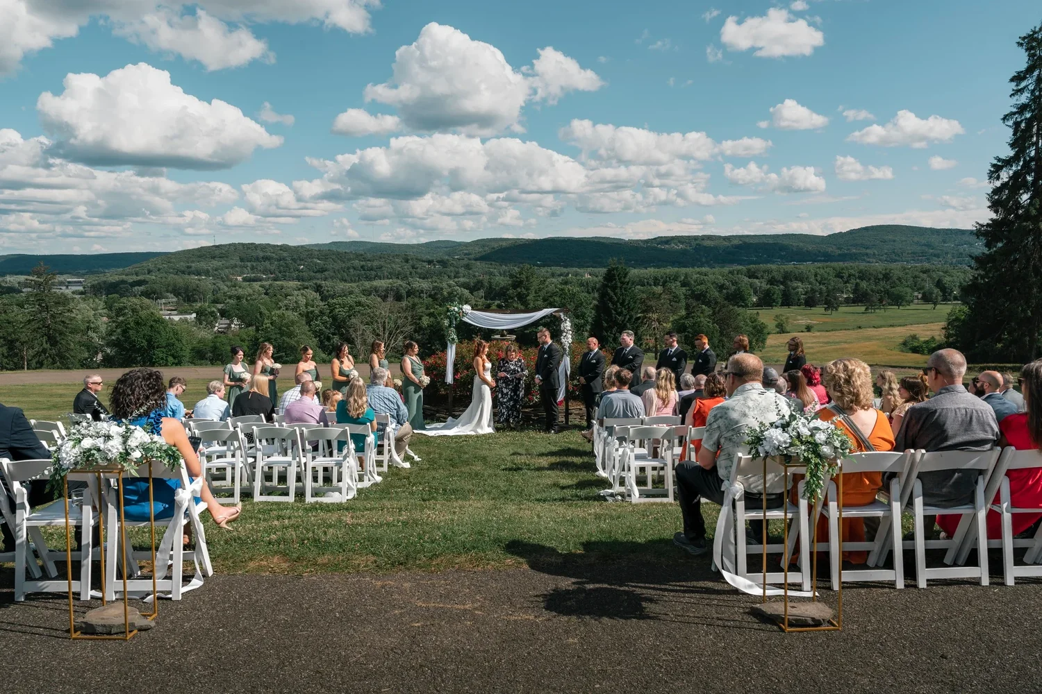 An outdoor wedding ceremony at Traditions at the Glen: guests in white chairs facing a draped wooden arbor, with the valley and hills beyond