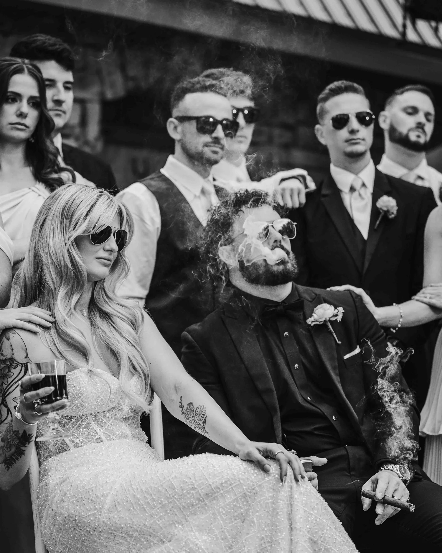 Black and white photograph of Kari and Josh seated in sunglasses with their wedding party behind them, Josh exhaling cigar smoke.