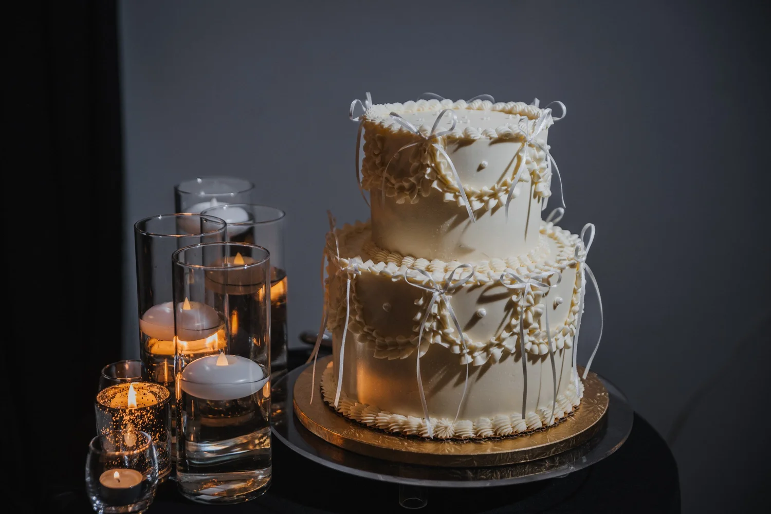 A two-tier white cake with piped details beside floating candles on a black table