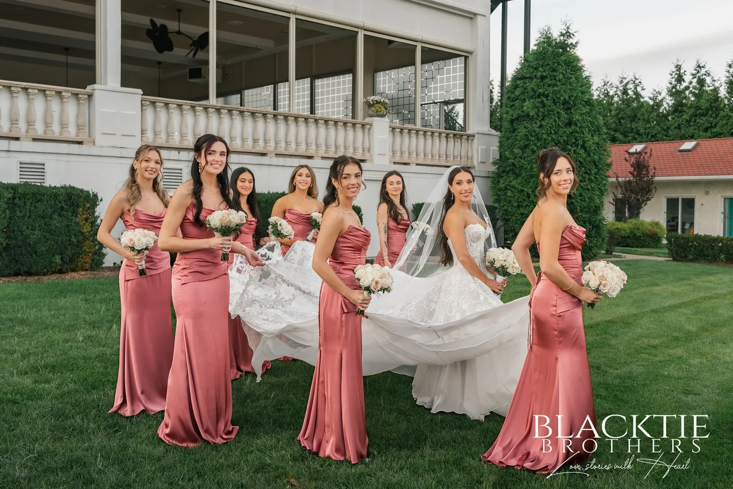 Bridesmaids in dusty rose satin gowns on the lawn, several of them lifting and spreading Leah's long veil as she stands among them, the white terrace railing and the building behind