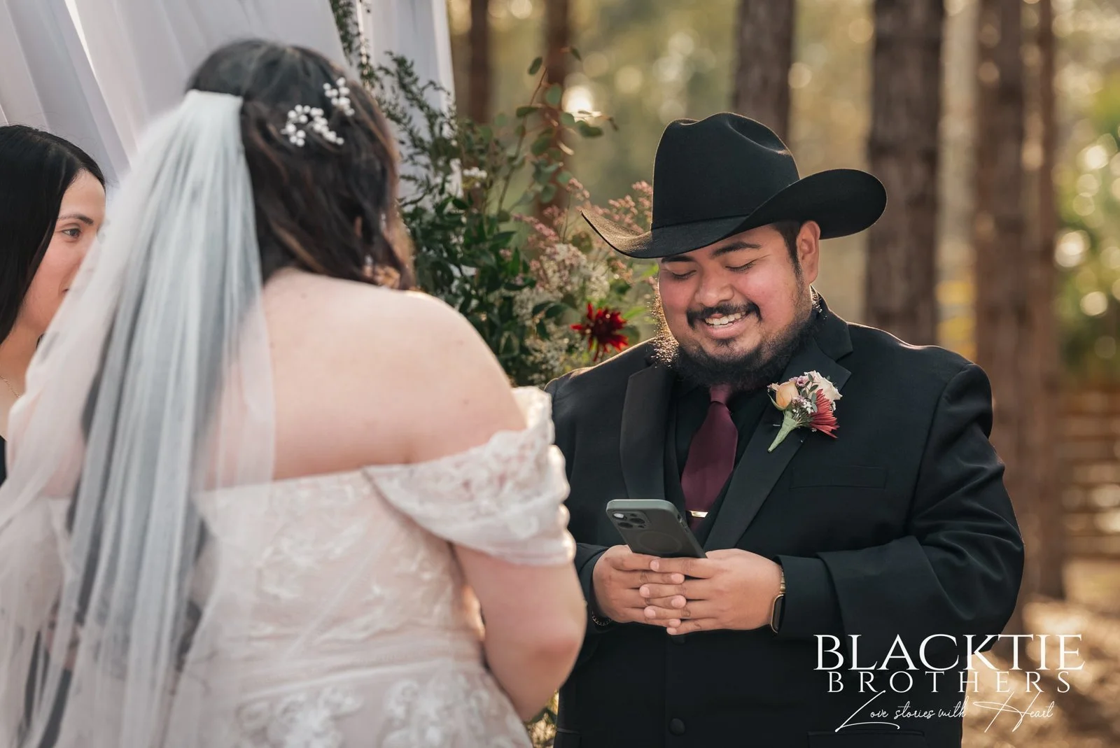 Alberto smiles while reading from his phone during the ceremony, facing Alyssa in her veil.