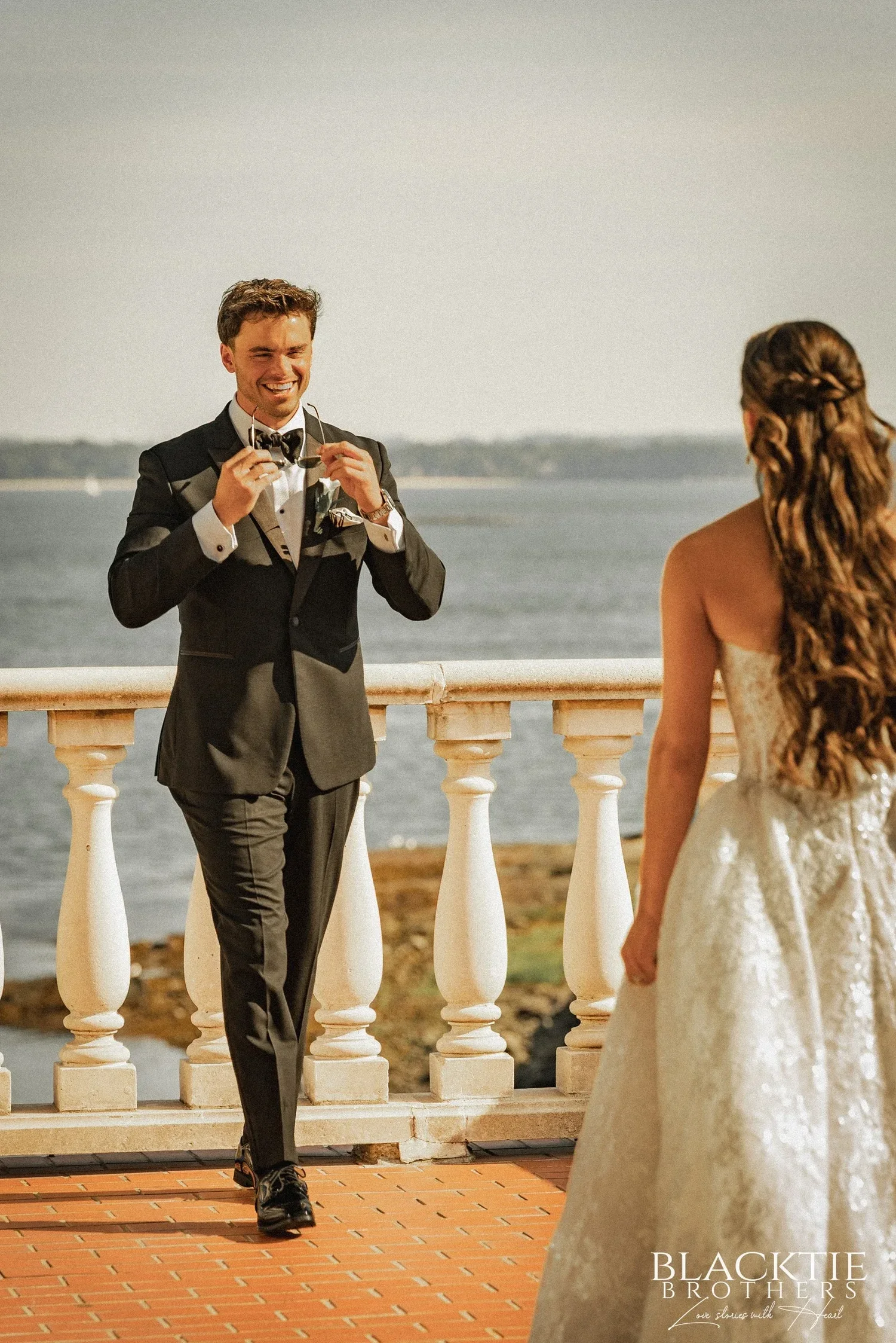 Nicholas turned around at the terrace balustrade, grinning with his hands raised to his face, as Leah stands in front of him with her back to the camera and the water behind