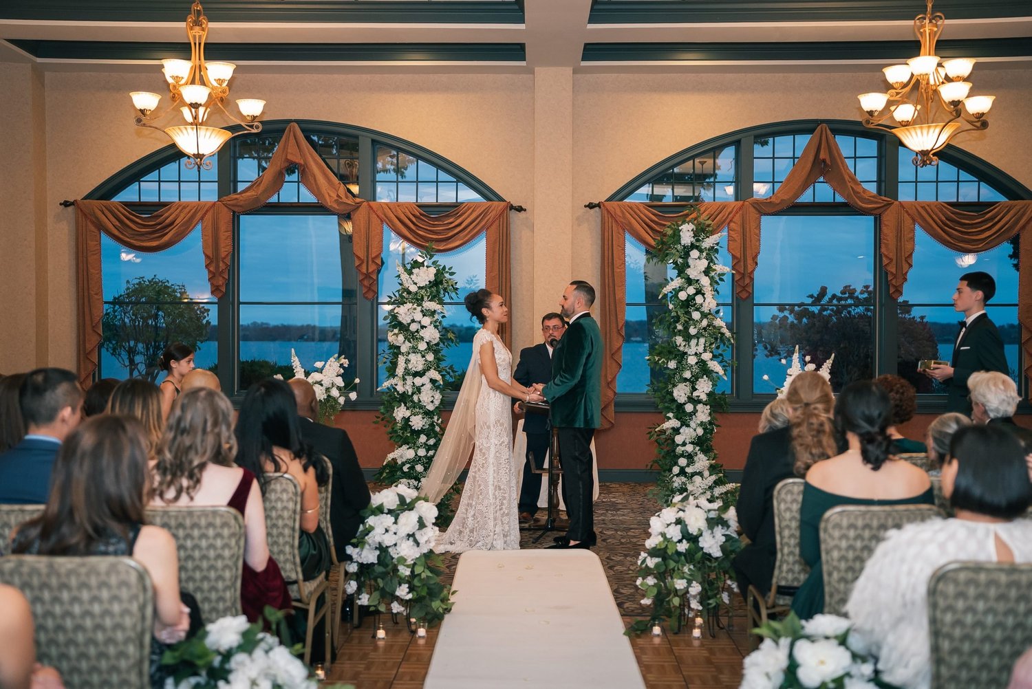Shannon and Leon hand in hand at the front of the Meritage Ballroom at Belhurst, with Seneca Lake in the arched windows behind them