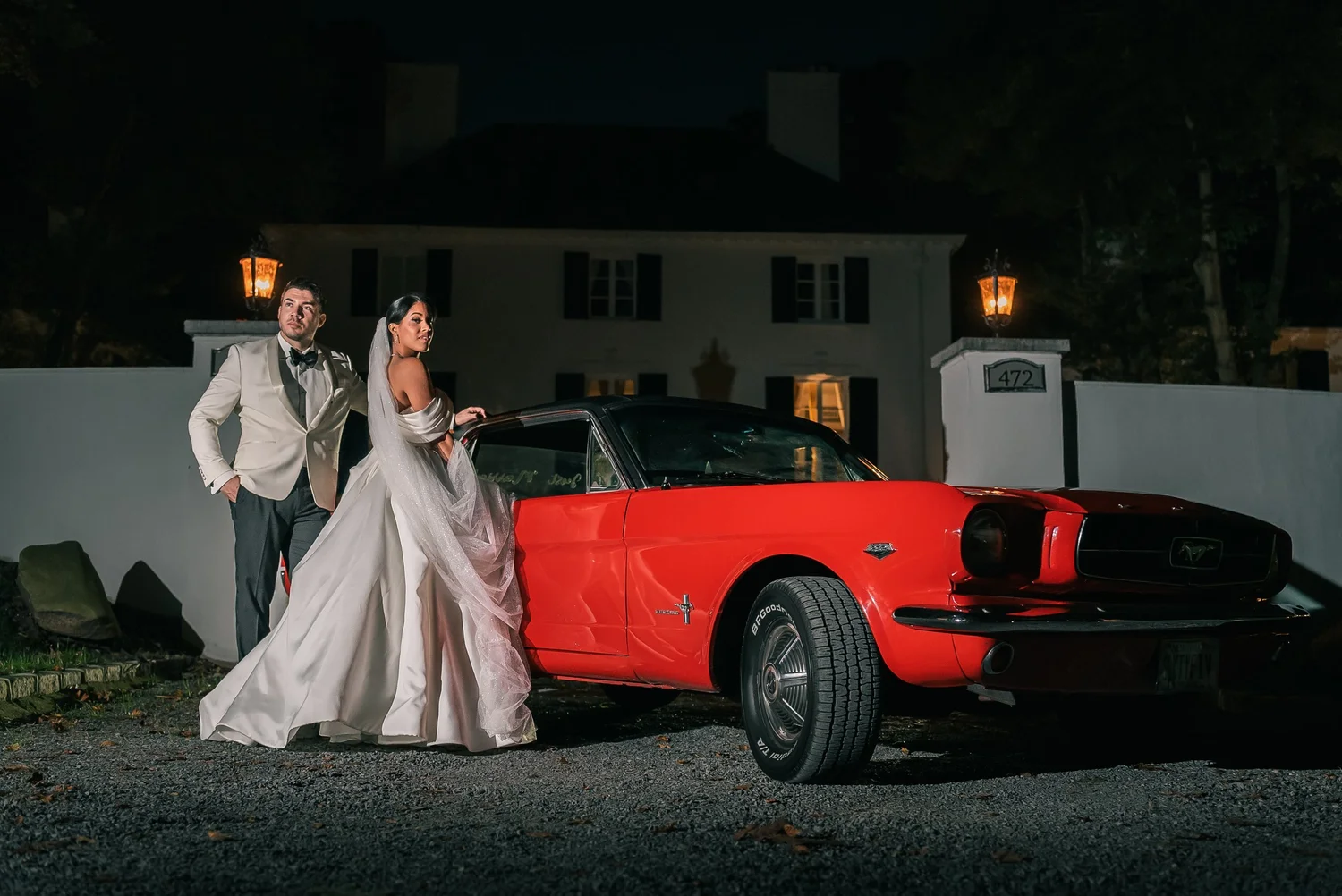 Patricia, her veil trailing, and Joshua stand beside a bright red vintage Mustang under lantern light at night.