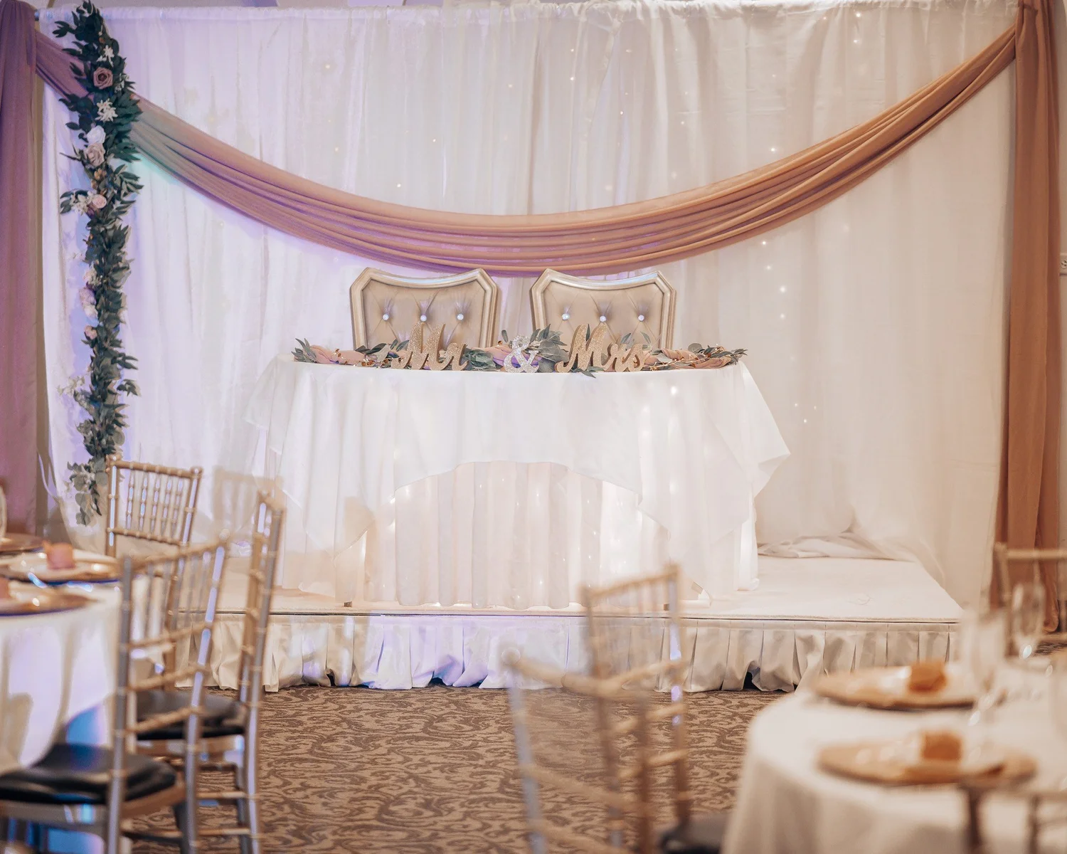 The head table in front of white and blush draped fabric with greenery, set before the reception