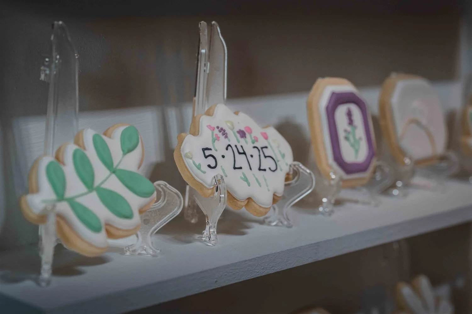 Decorated wedding cookies displayed on clear stands, including one iced with the date 5-24-25.