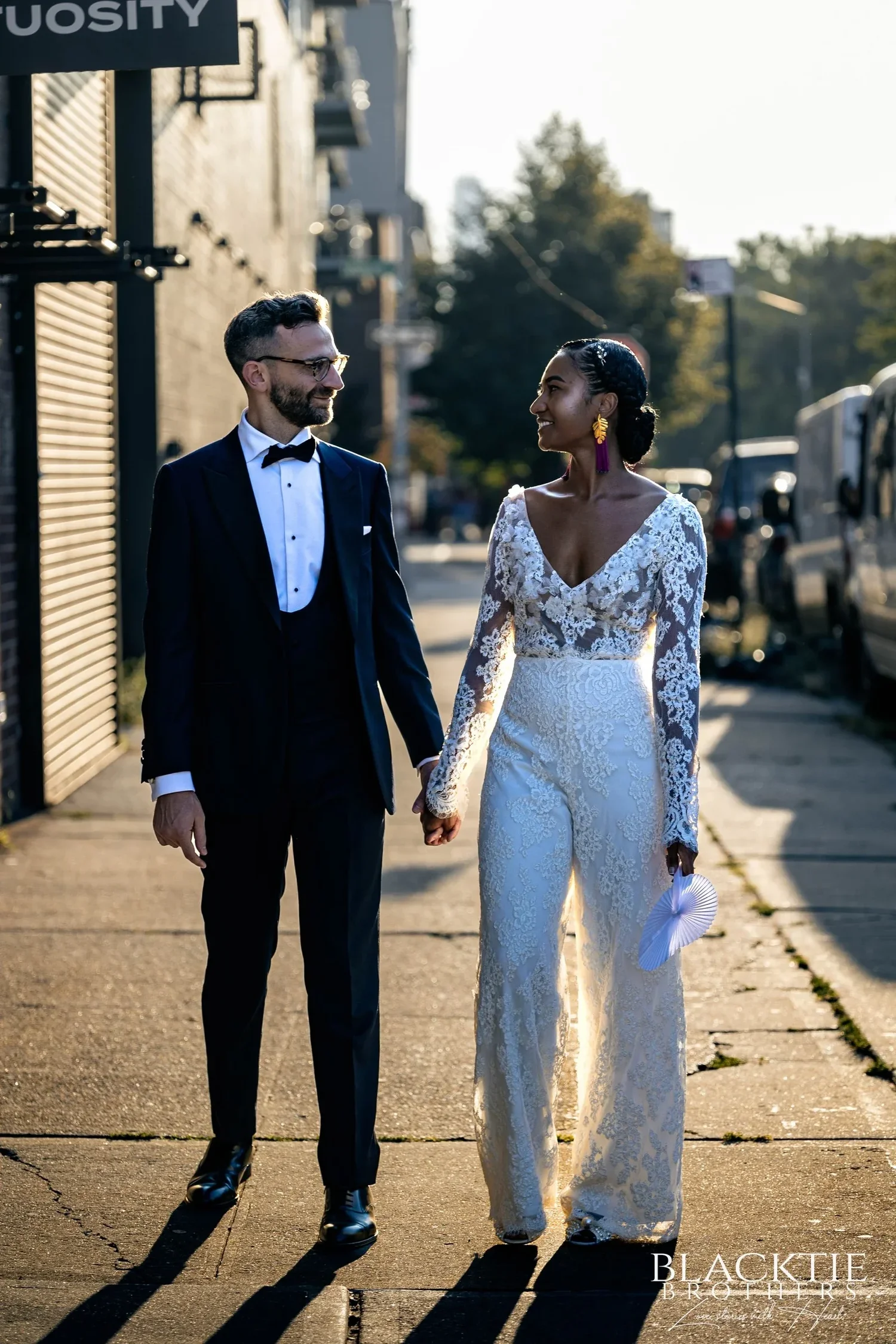 Ciara and Drew holding hands on a sunlit Brooklyn sidewalk, long shadows at their feet, roll-down shutters and trees along the street