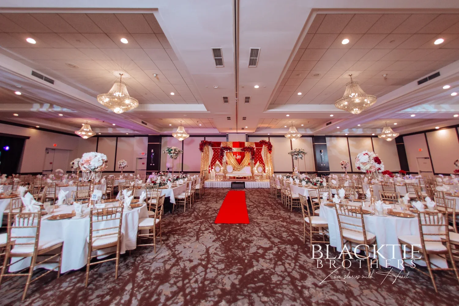 A large ballroom at the DoubleTree by Hilton Binghamton set with round tables and gold chairs under crystal chandeliers, a red runner leading to a red and gold wedding stage
